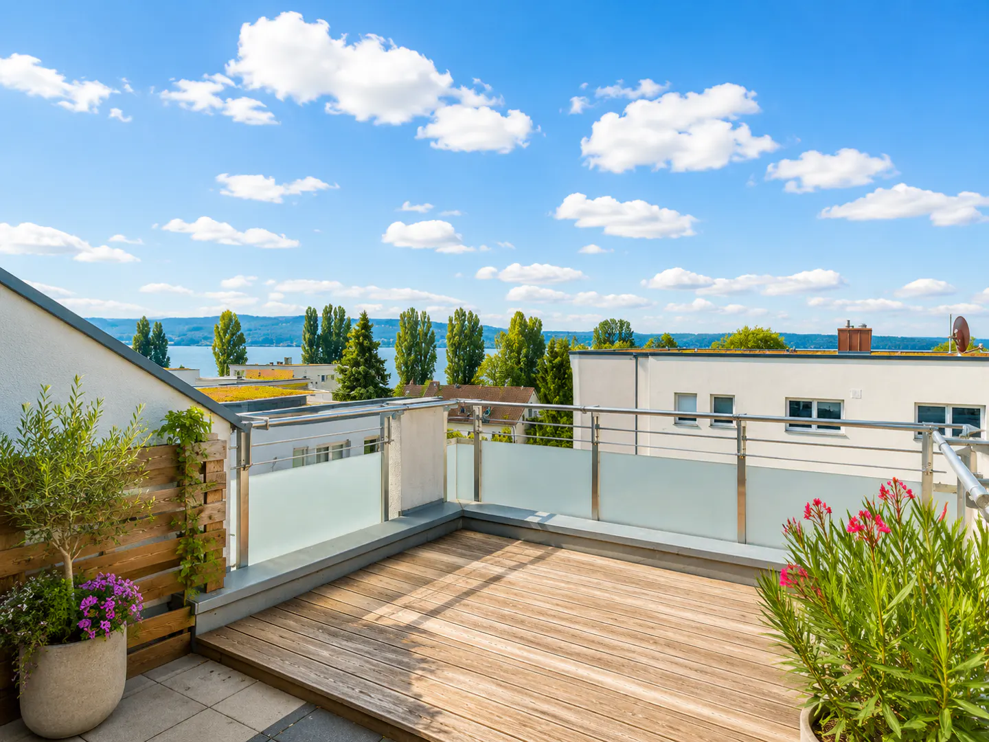 Sunny rooftop terrace with wooden decking, glass railings, potted plants, and a scenic view of a lake and blue sky.
