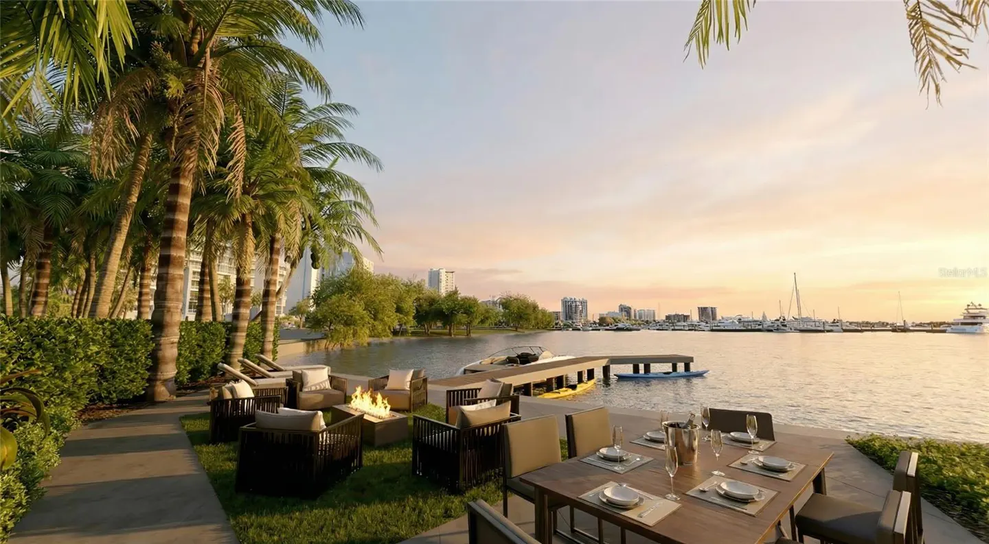 Outdoor patio with dining table, chairs, and fire pit overlooking a bay with boats and a city skyline at sunset. Palm trees line the property.