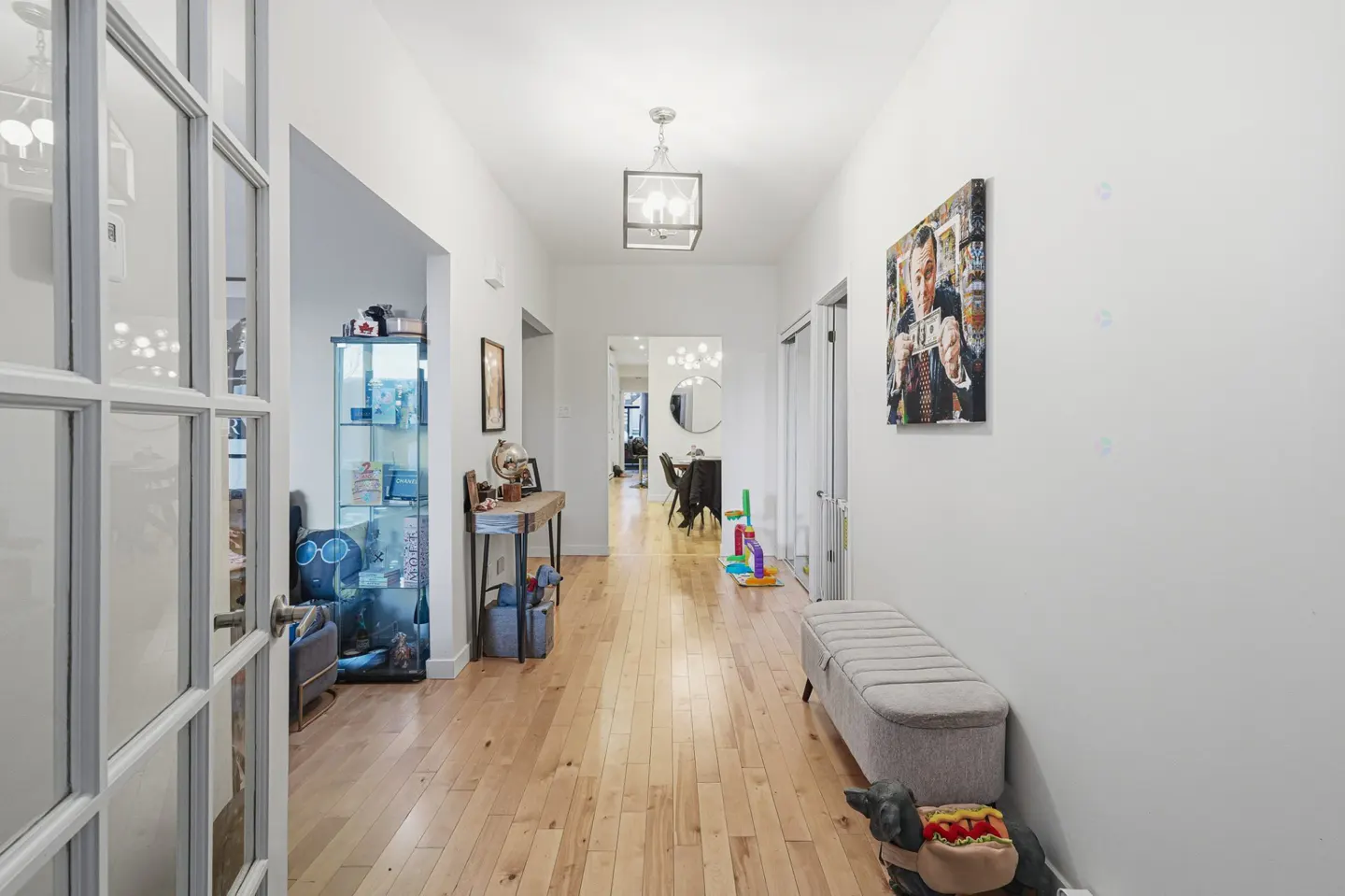 Hallway view with light wood floors, white walls, and a gray bench. A framed picture hangs on the wall. A glass cabinet is visible through a doorway.