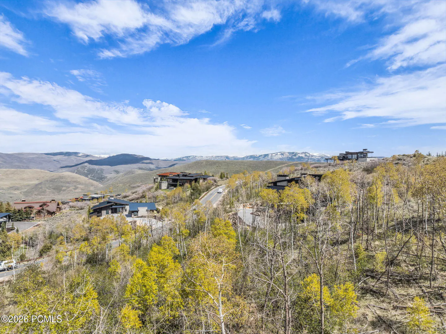 Scenic view of luxury homes nestled in a hillside with autumn trees under a blue, cloudy sky. Mountains are visible in the distance.
