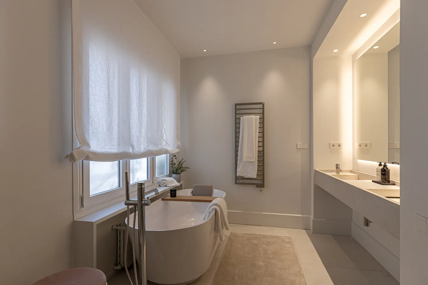Modern bathroom with a white freestanding tub, a double vanity with a large mirror, and a heated towel rack on the wall.