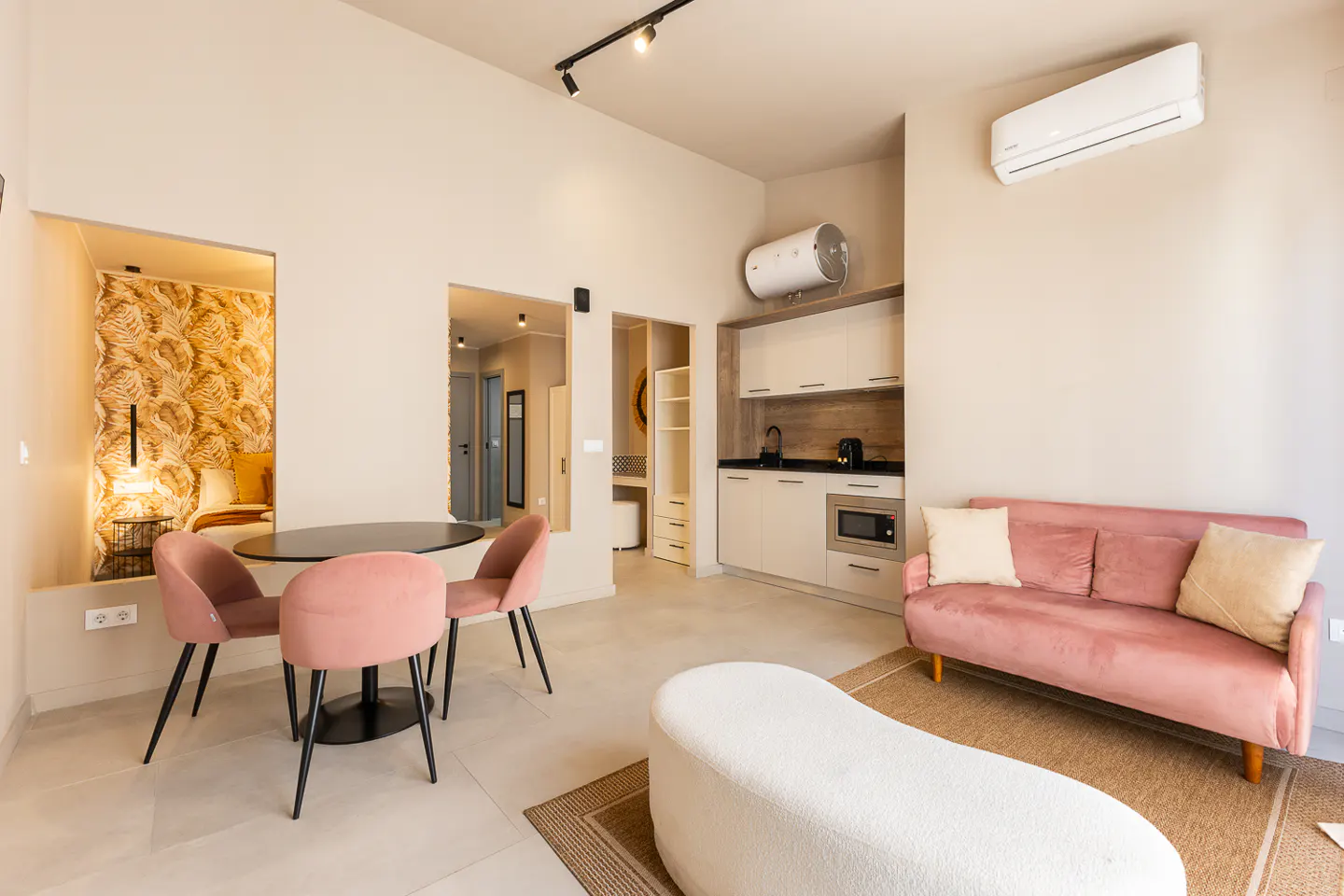 A modern apartment interior with a pink sofa, round table with pink chairs, and a kitchenette. A bedroom nook is visible in the background.