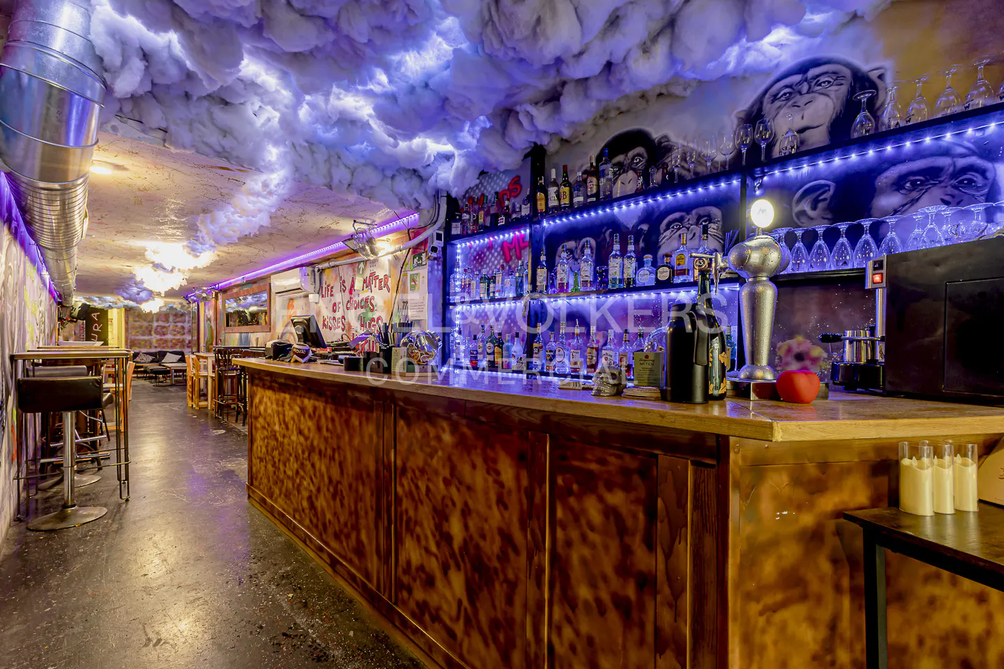 Interior view of a bar with a wooden counter, shelves of liquor, monkey murals, and cloud-like ceiling decor with blue lighting.