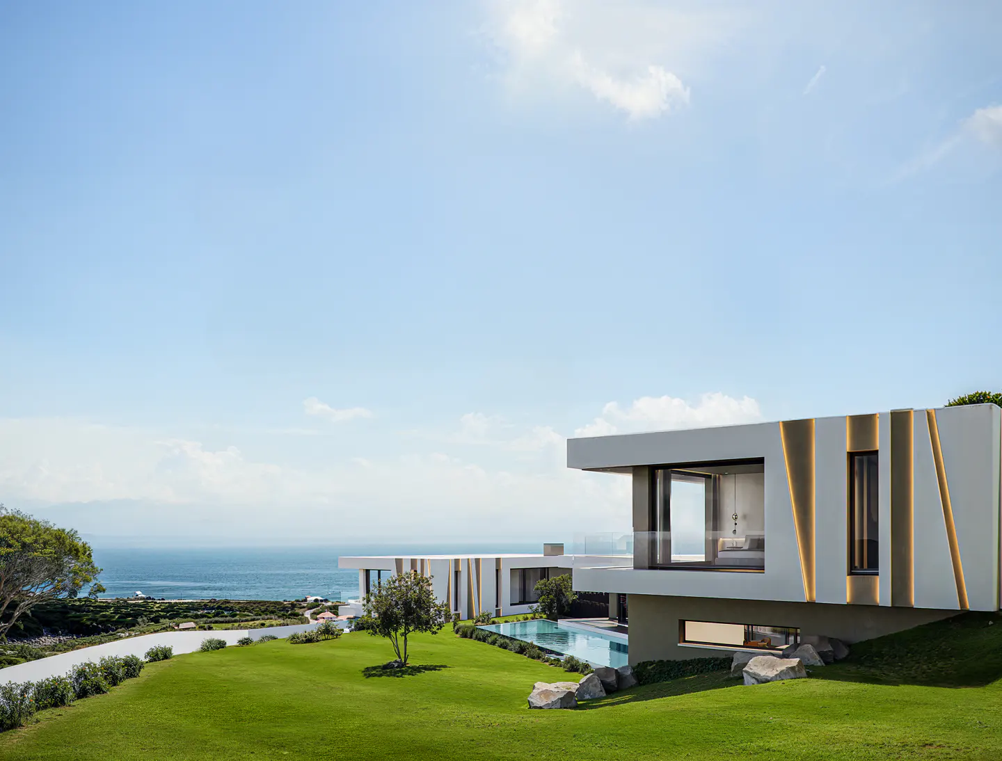 Modern white house with a pool on a green lawn overlooking the ocean under a blue sky.