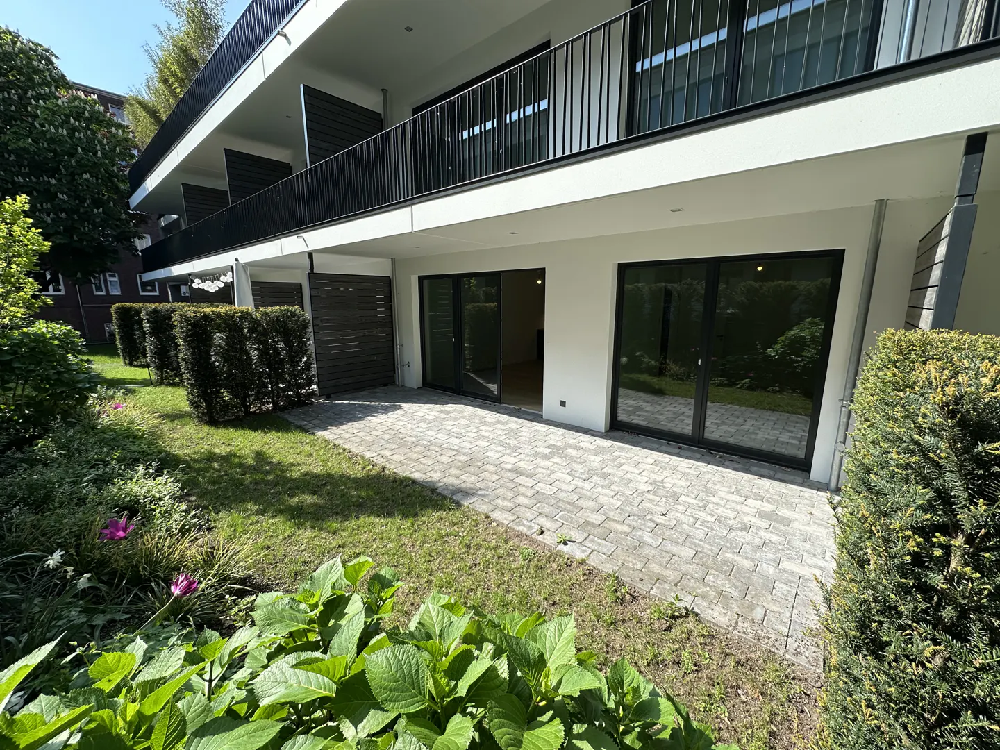 Exterior view of a modern two-story apartment building with a patio and garden. The building is white with black railings and window frames.