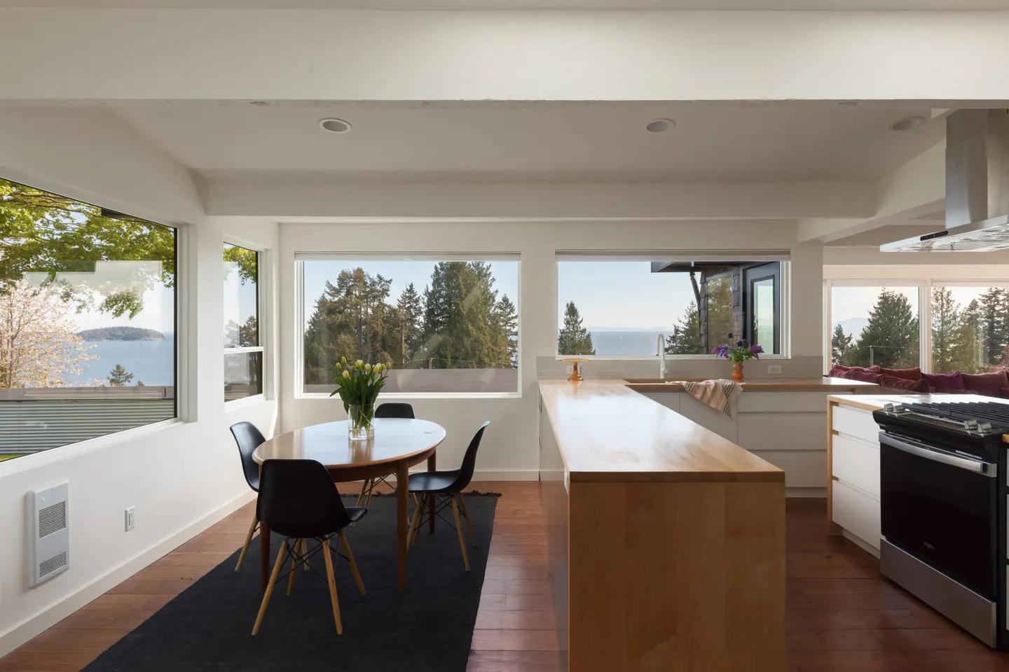 Bright, modern kitchen with wood floors, white walls, and large windows with ocean views. A dining table with black chairs sits on a dark rug.