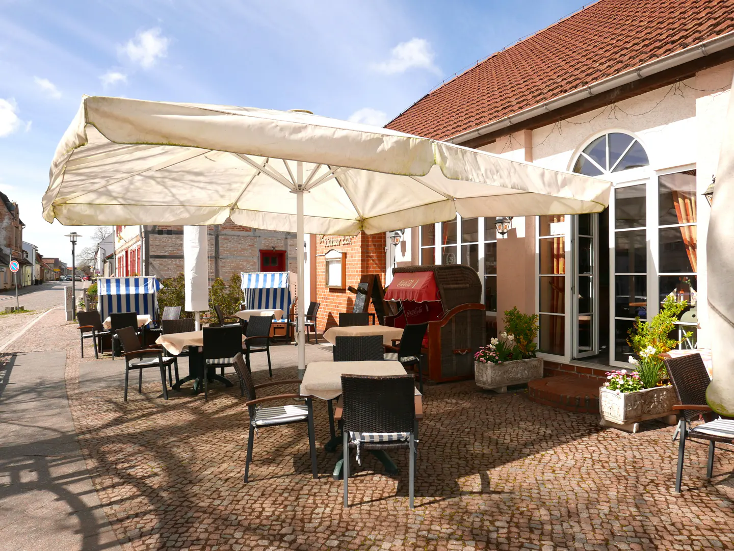 Outdoor cafe patio with tables, chairs, and large white umbrellas on a cobblestone terrace next to a brick building.