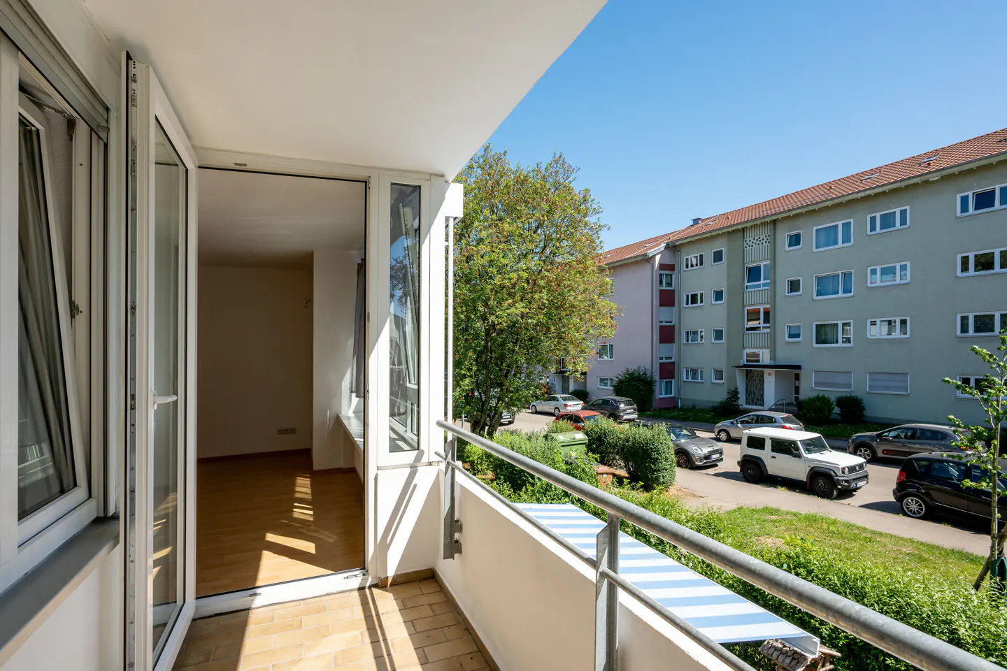 View from a balcony looking into an empty room with wood floors and out toward a street with parked cars and apartment buildings.
