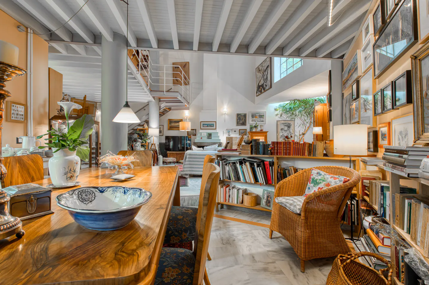 Open-concept living space with a long wooden table, wicker chair, and bookshelves filled with books and art. White walls and ceiling beams.