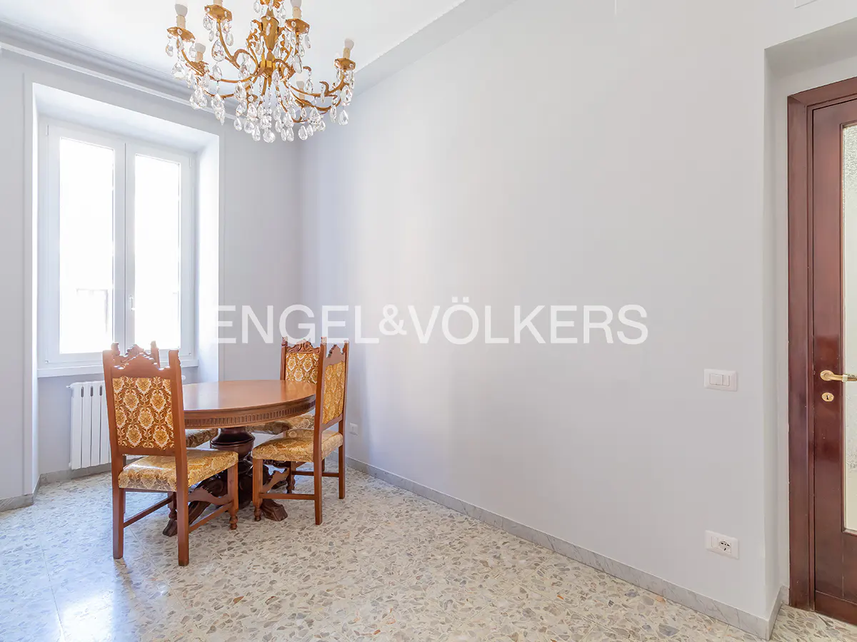 Dining area with a round wooden table, two patterned chairs, a crystal chandelier, and terrazzo flooring.