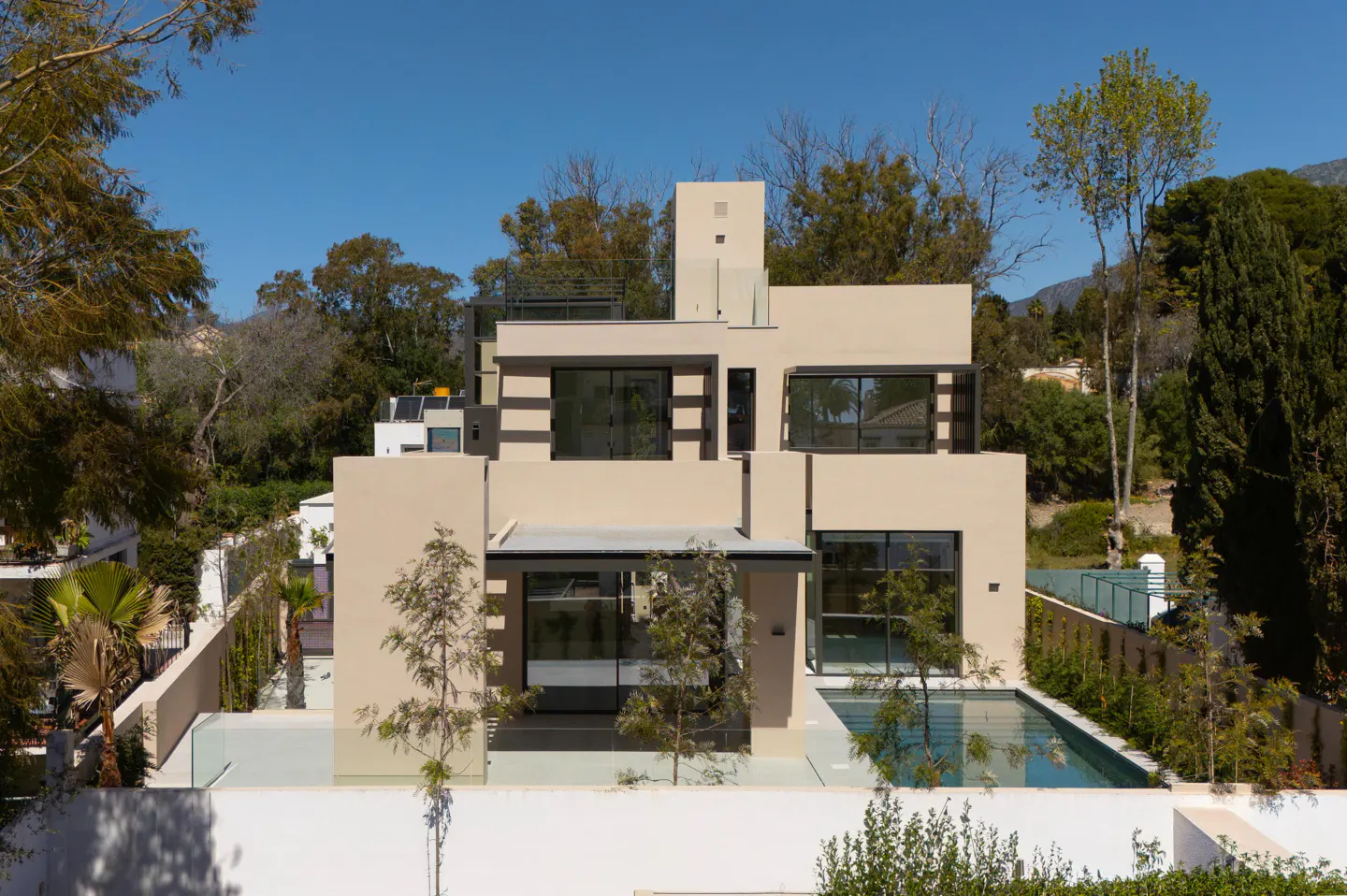Modern beige house with black-framed windows, a pool, and green trees against a clear blue sky.