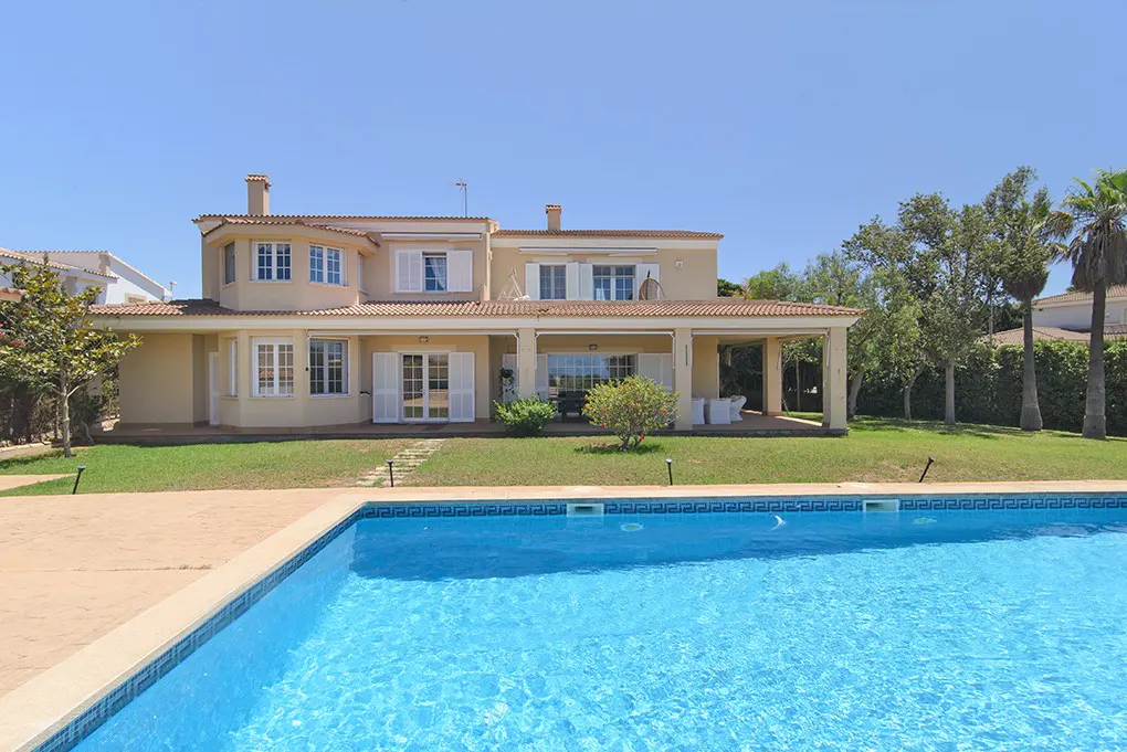 A two-story beige villa with a covered patio, green lawn, and a bright blue swimming pool under a clear sky.
