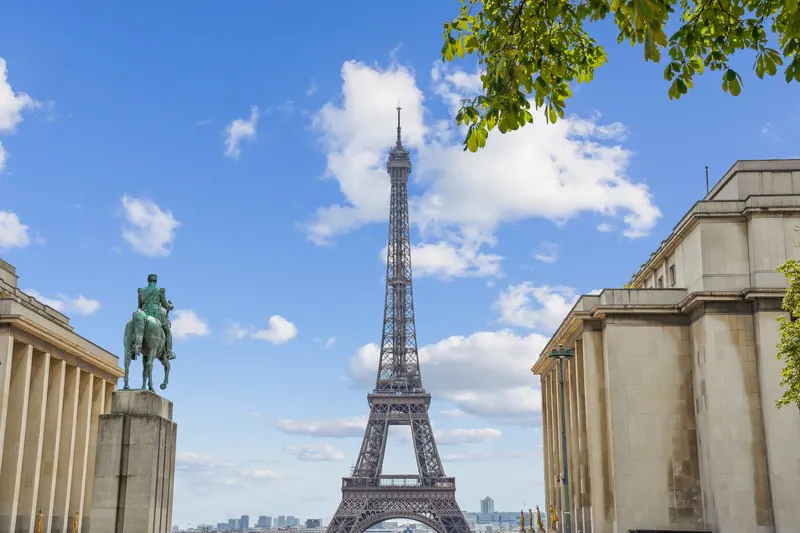 Eiffel Tower view from Trocadéro, Paris. Blue sky with clouds, statue of man on horse, and tan buildings.
