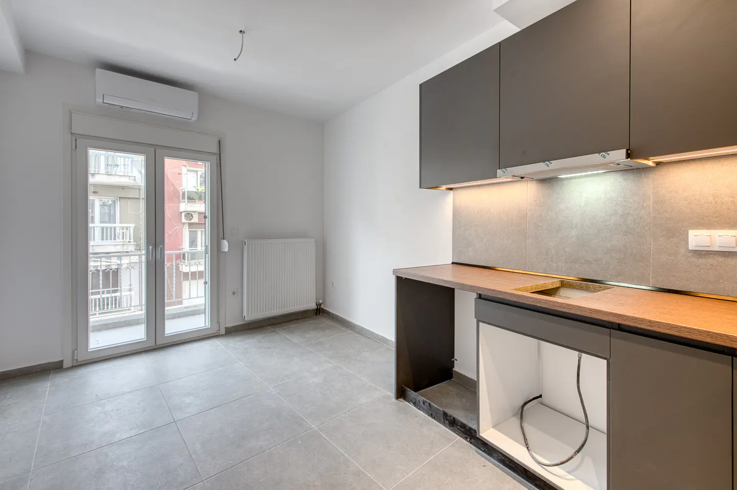 A bright, modern kitchen with gray cabinets, wood countertop, and gray tile flooring. A double glass door leads to a balcony.