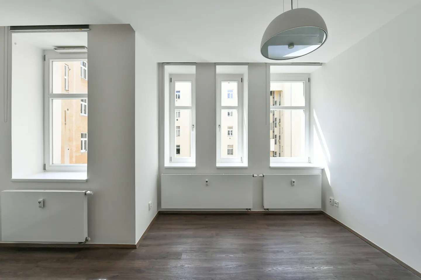 Bright, empty room with white walls, dark wood floors, four windows, white radiators, and a modern dome pendant light.