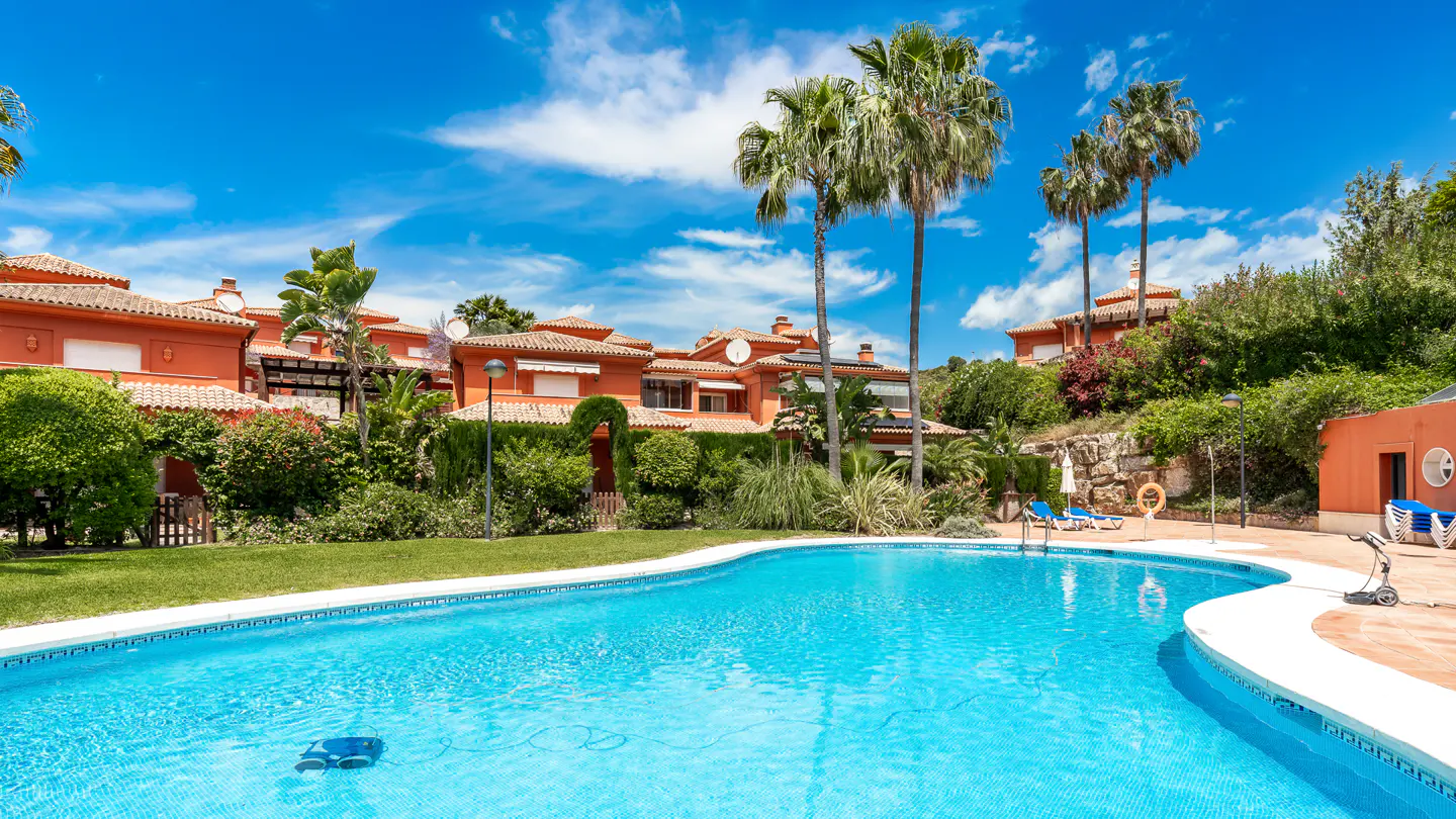 A turquoise pool in front of orange townhouses with palm trees under a blue, cloudy sky.