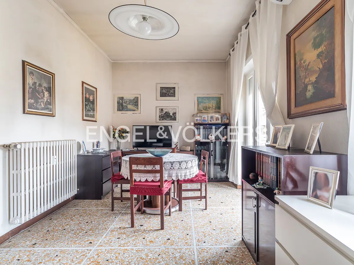 A bright dining room with a round table, red chairs, and a lace tablecloth. Artwork adorns the walls, and a radiator sits near a dark cabinet.