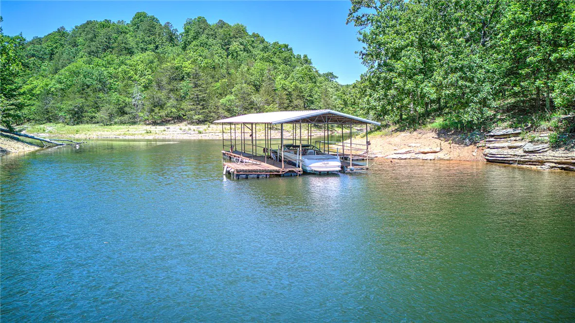 A boat is docked under a metal awning on a lake surrounded by green trees.