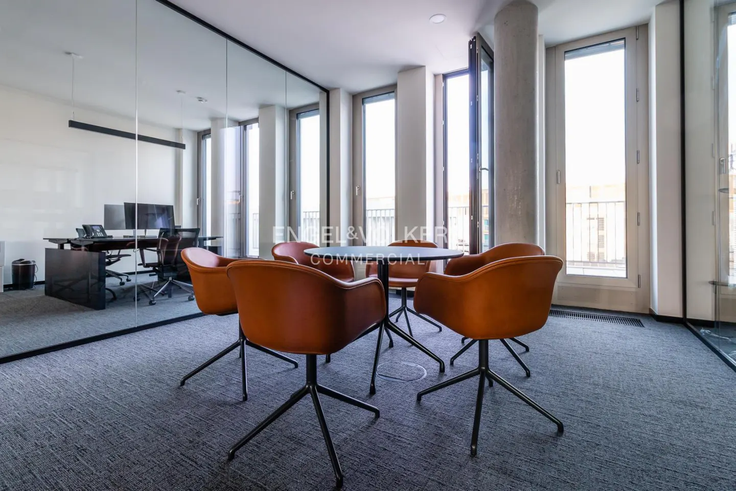 Office space with a round table surrounded by six brown chairs on a gray carpet, near a glass-walled office.