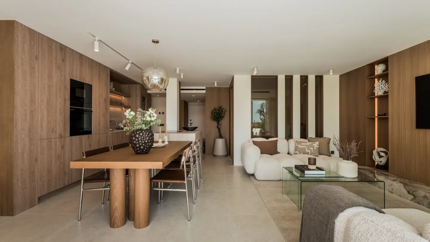 Modern open-plan living area with a wooden dining table, white sofa, glass coffee table, and warm wood wall paneling.