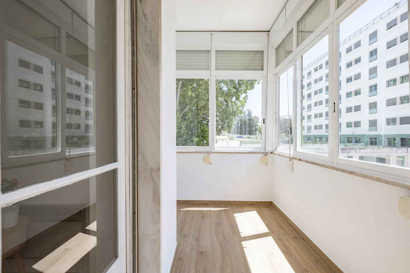 Bright, enclosed balcony with light wood flooring, white walls, and large windows overlooking a neighboring apartment block.