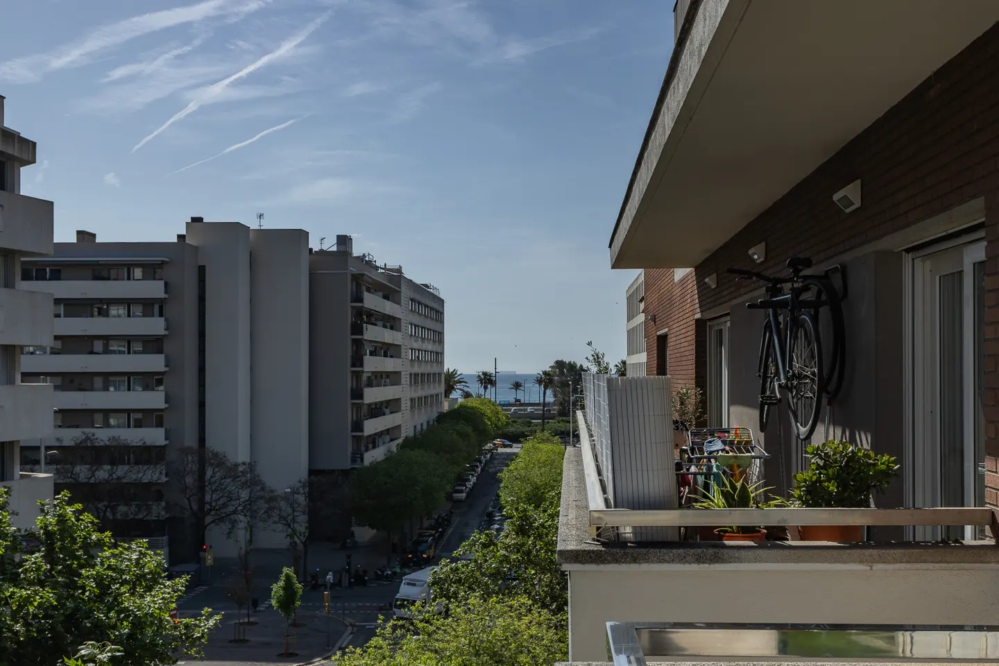 View from a balcony with a bicycle hanging on the wall, overlooking a street lined with trees and buildings, leading to the sea.
