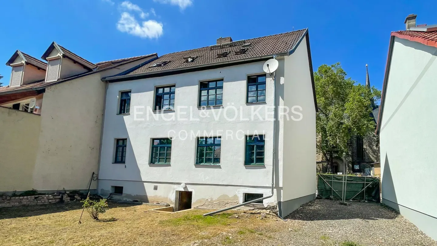 Exterior view of a three-story white building with dark green window frames and a brown roof on a sunny day.