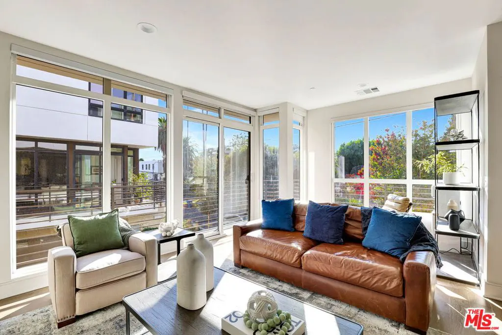 Bright living room with a brown leather sofa, blue pillows, and large windows showing a sunny outdoor view.