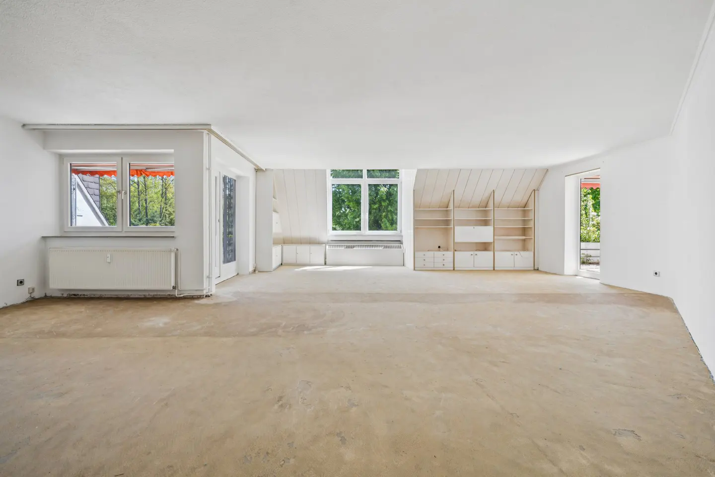 Bright, empty room with white walls, large windows, and built-in shelving. The floor is bare, and natural light streams in.