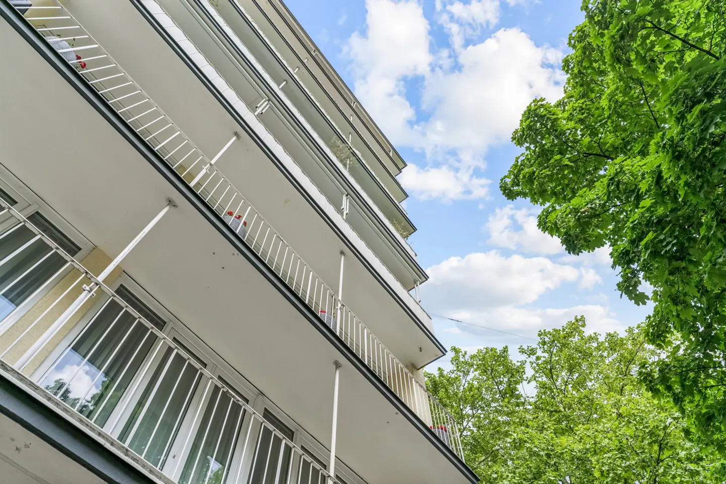 Low angle view of a white apartment building with balconies, green trees, and a blue sky with clouds.