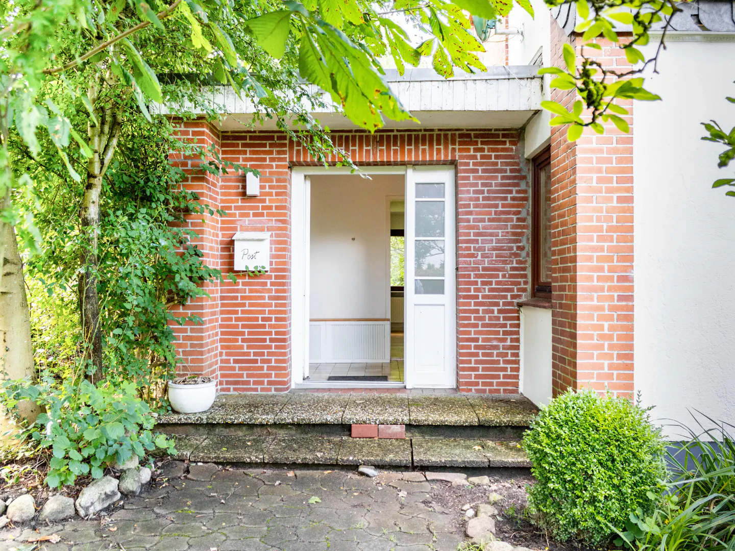 Exterior view of a brick house entrance with an open white door, a mailbox, and green foliage.