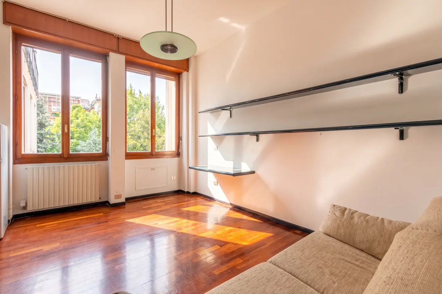 Bright living room with hardwood floors, large windows, and a beige sofa. Black shelves are mounted on the white wall.