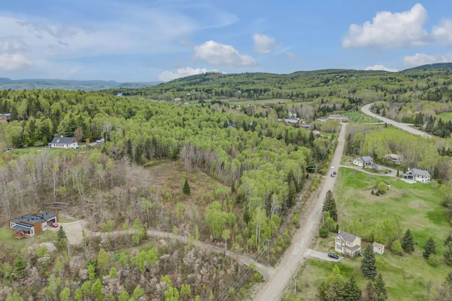 Aerial view of a rural landscape with scattered houses, a dirt road, and dense green forests under a blue, cloudy sky.