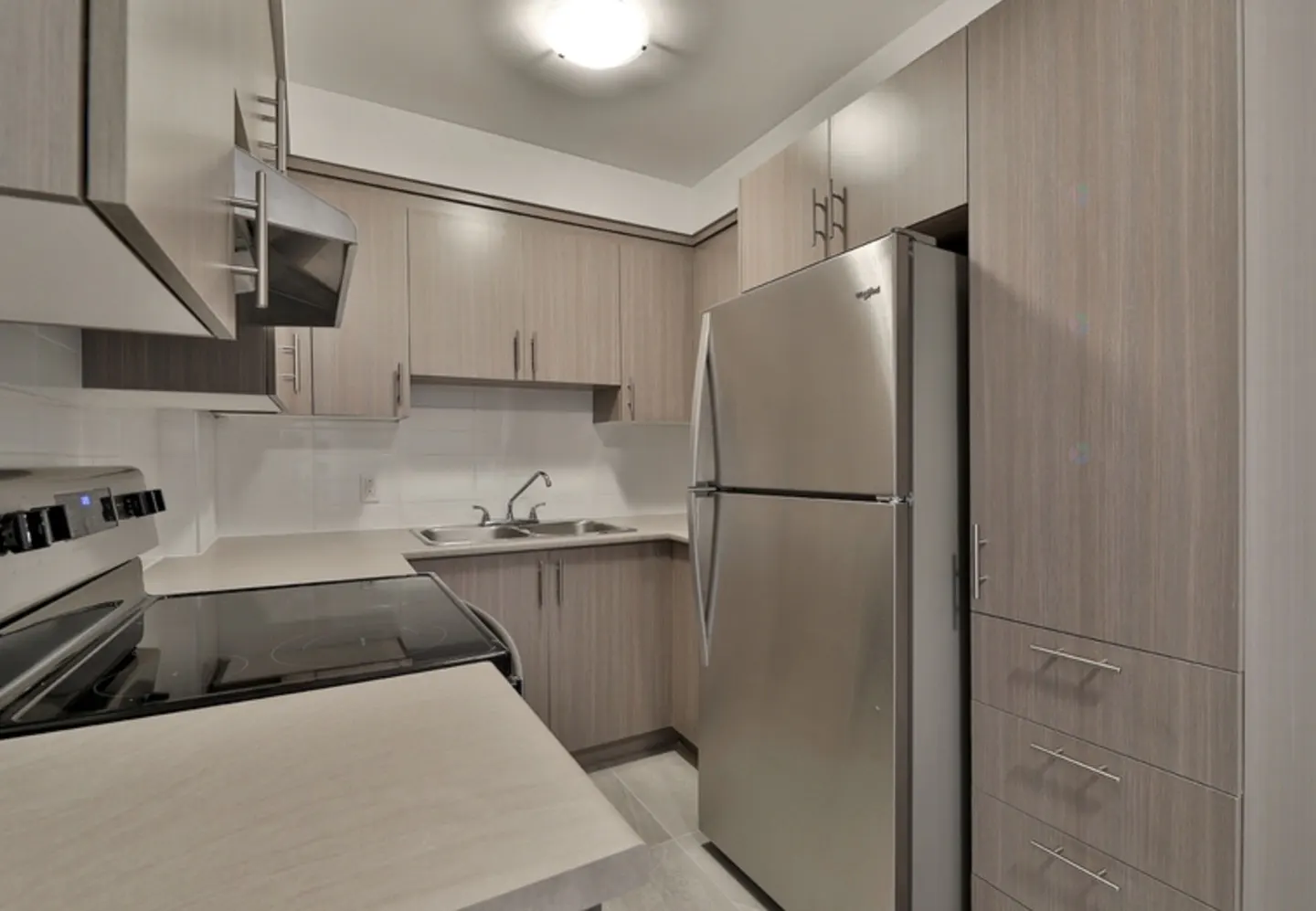 Modern kitchen with light wood cabinets, stainless steel appliances, and a double sink under a ceiling light.