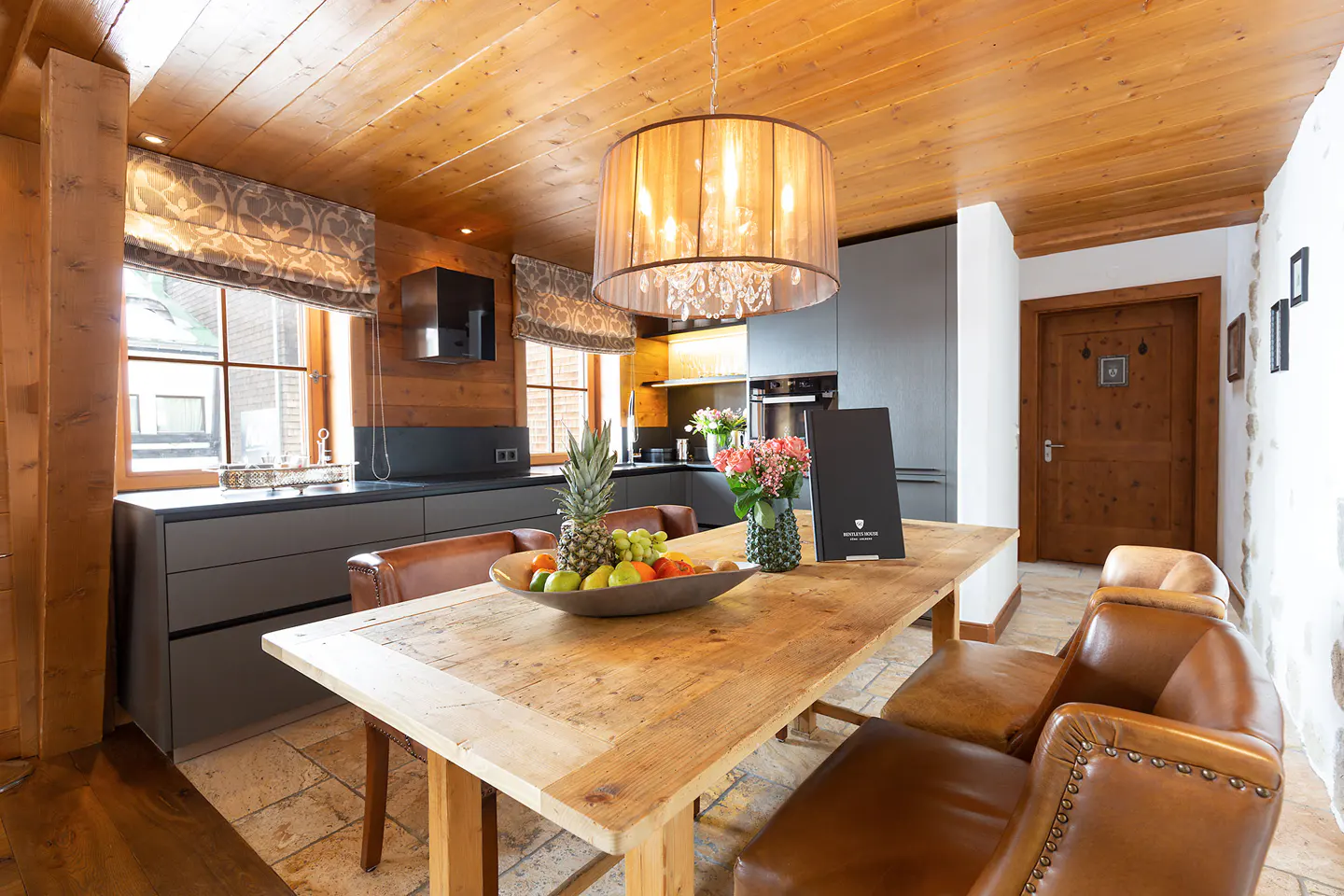 A rustic kitchen with a wooden table, leather chairs, and a fruit bowl. The ceiling and walls are wood paneled, and a modern kitchen is in the background.