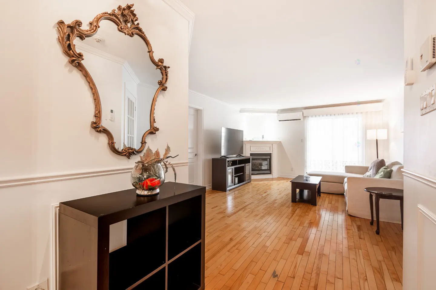 Bright living room with hardwood floors, white walls, and a large ornate mirror above a dark wood console table.