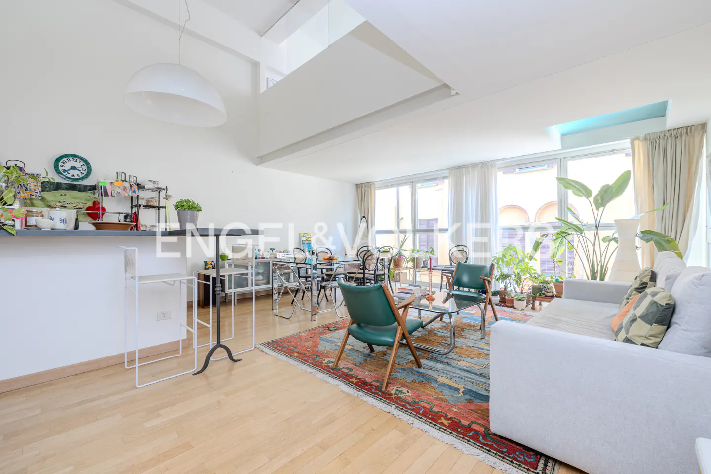 Bright living room with white walls, hardwood floors, a colorful rug, green armchairs, and large windows with plants.