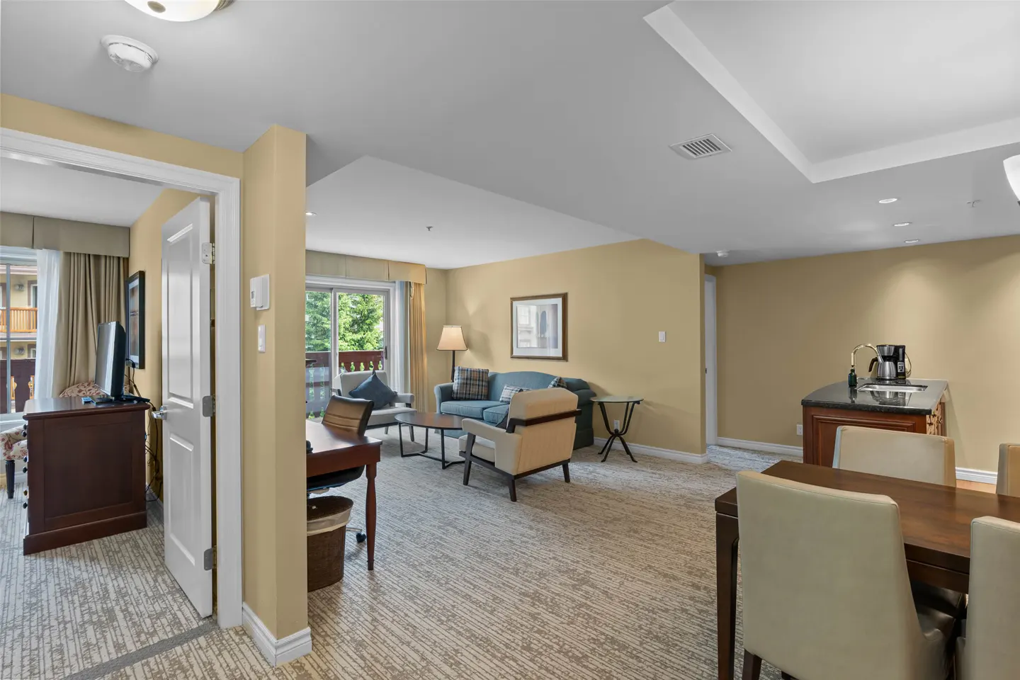 A wide shot of a hotel suite with a living room, dining area, and desk. The walls are tan, and the carpet is patterned. A blue sofa sits in the living room.