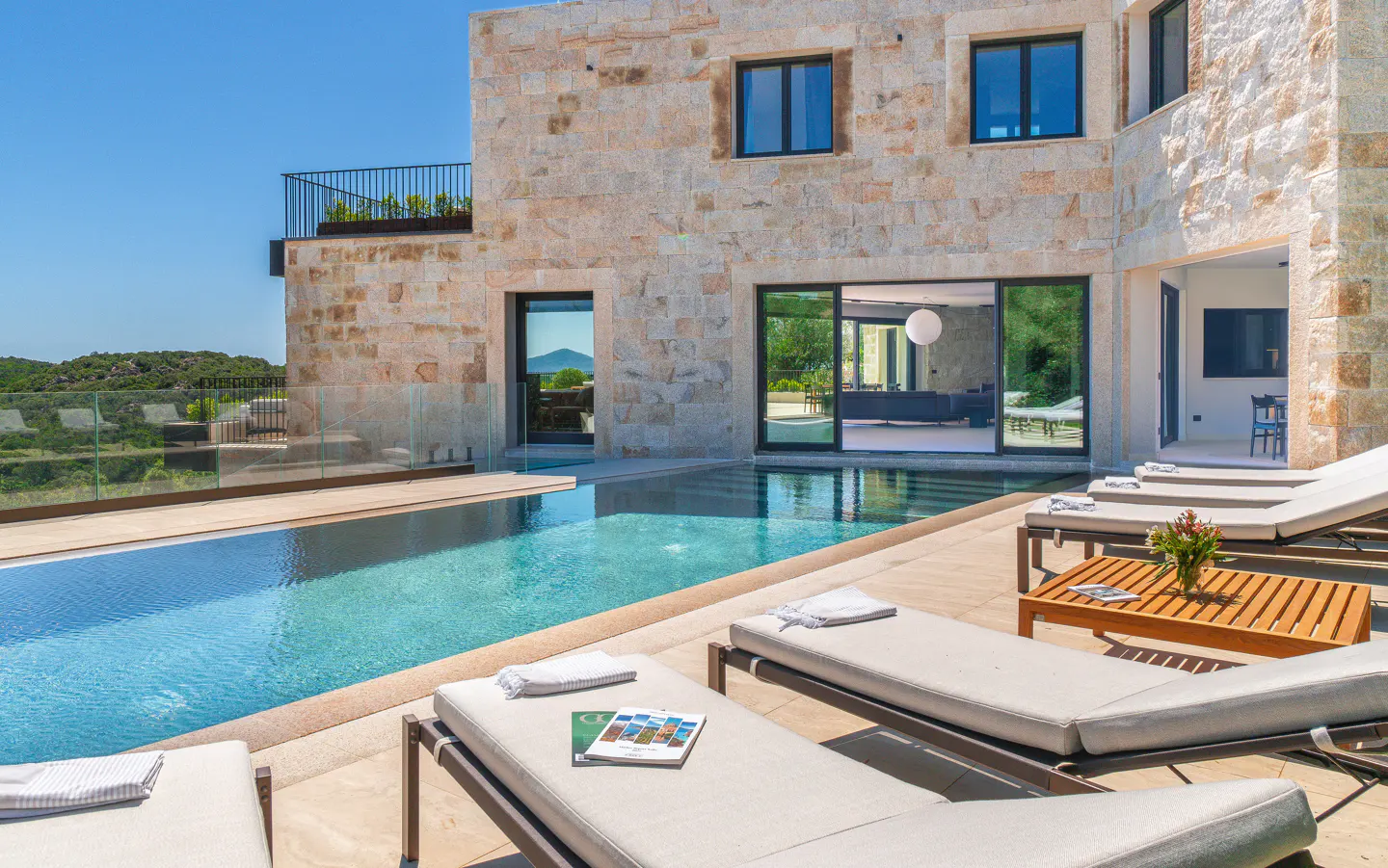 Modern stone villa with a swimming pool, lounge chairs on a patio, and a view of the surrounding landscape.