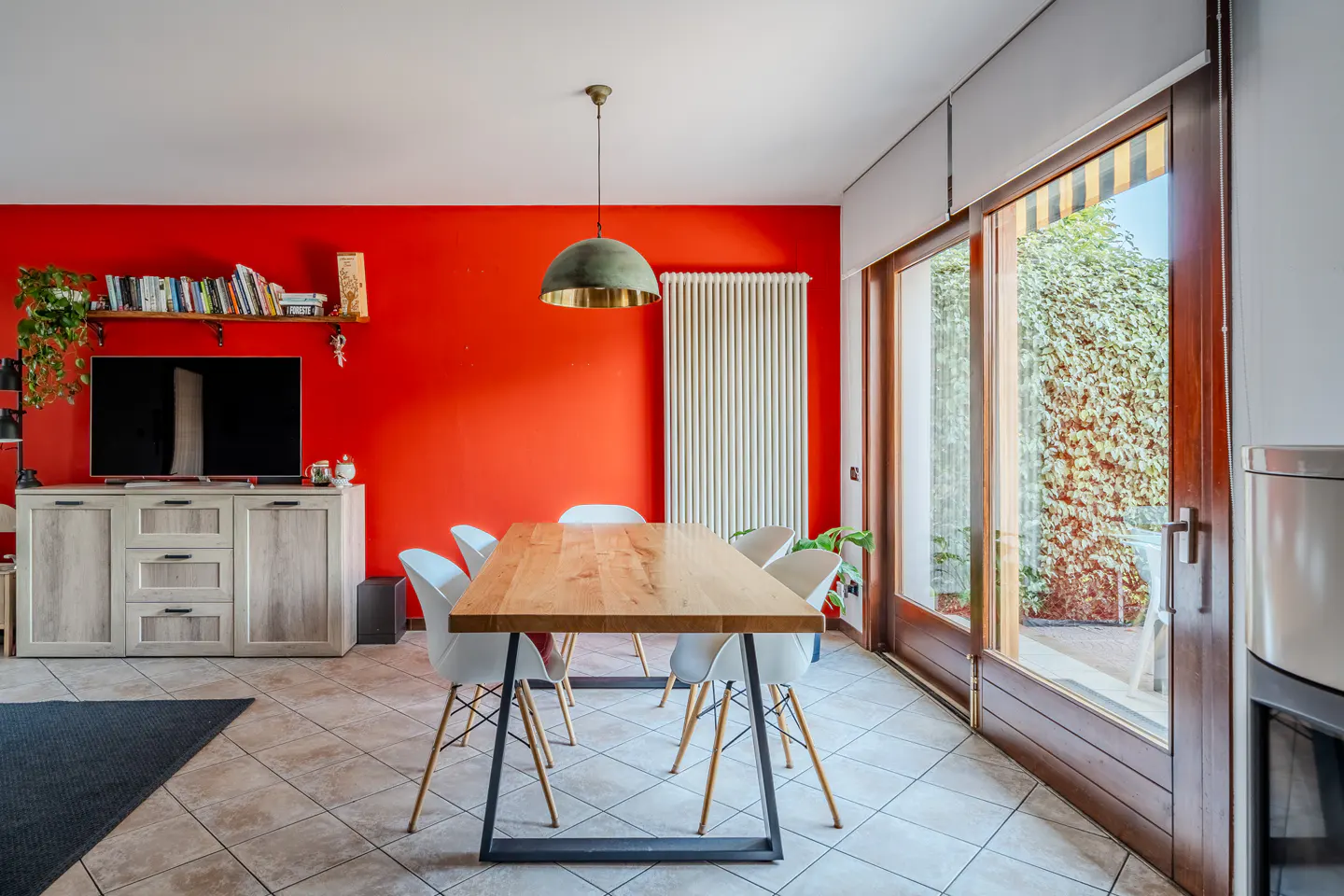A modern dining room with a wooden table, white chairs, and a red accent wall. Sliding glass doors lead to a patio with greenery.