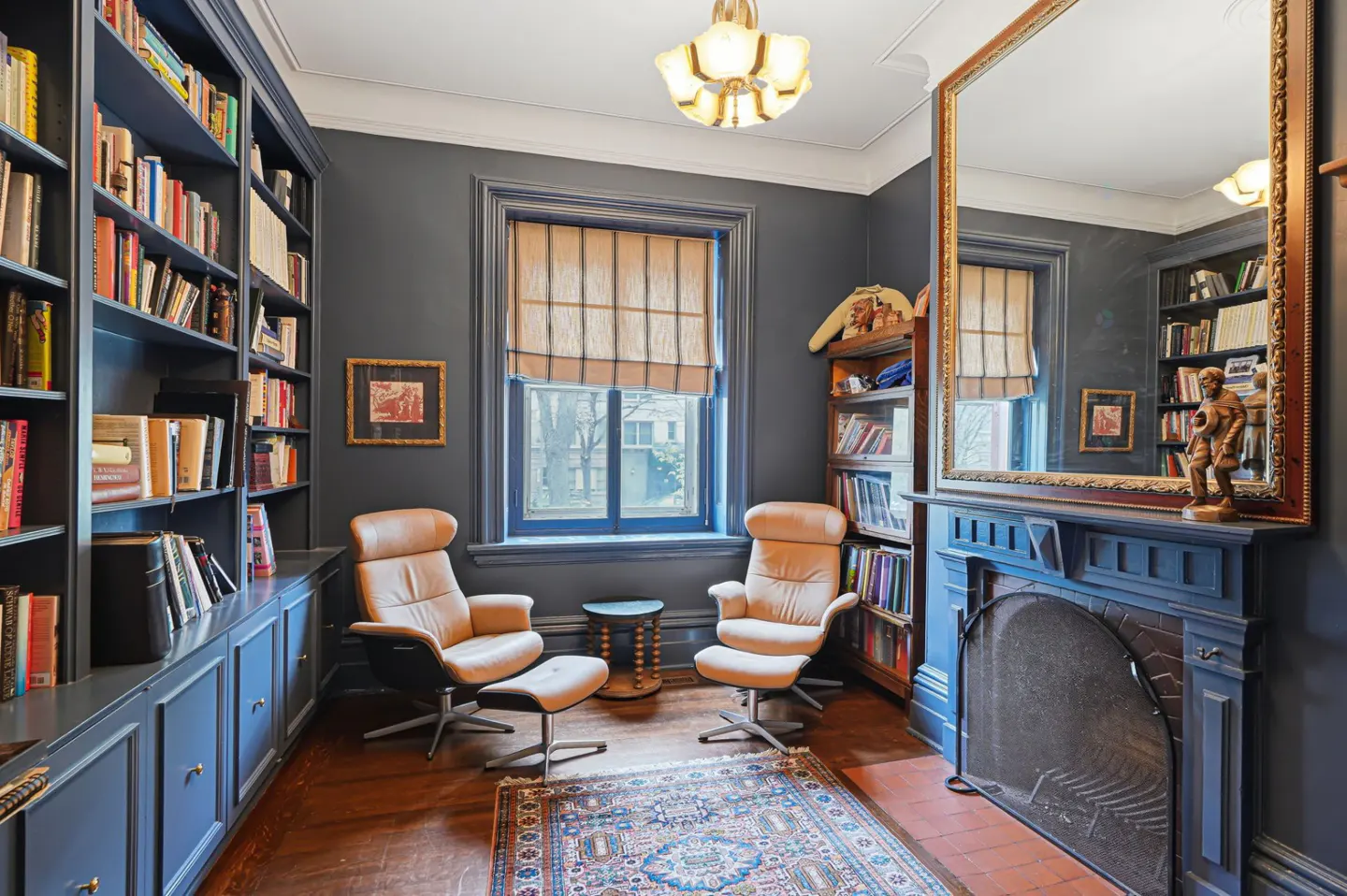 A cozy library with blue walls, filled bookshelves, and two tan leather chairs facing a window. A fireplace with a large mirror above it adds to the room's charm.