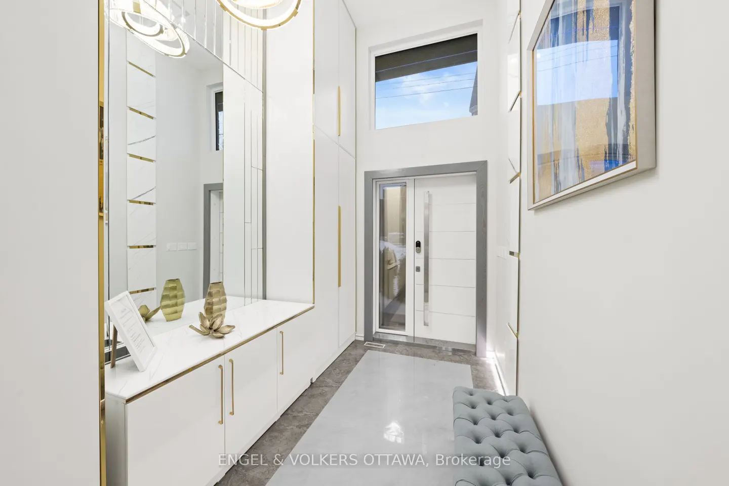 Bright foyer with white walls, cabinets, and a gray floor. A tufted bench sits near the front door, with a mirror and artwork adding visual interest.