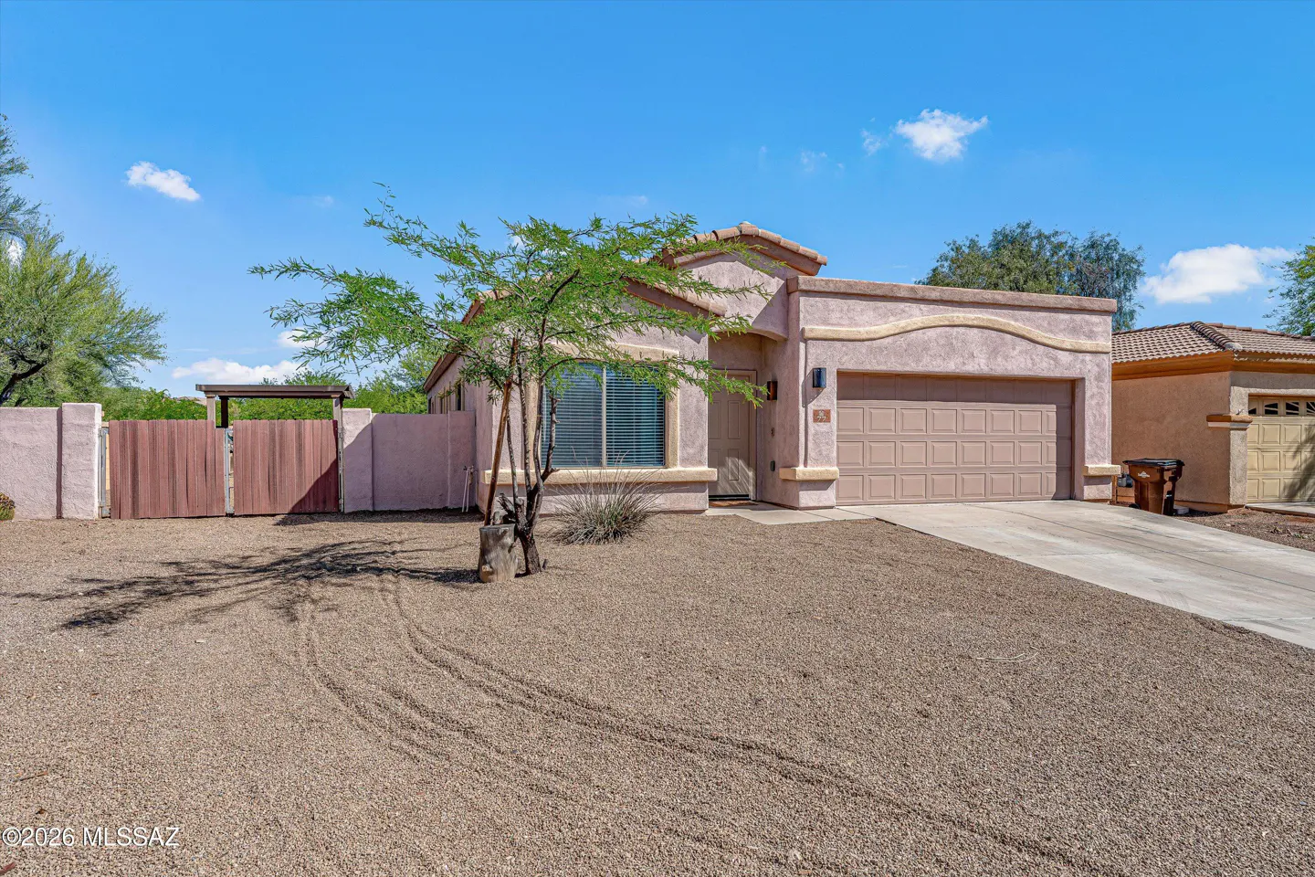 Tan stucco house with a brown garage door, a small tree, and a gravel yard under a blue sky.