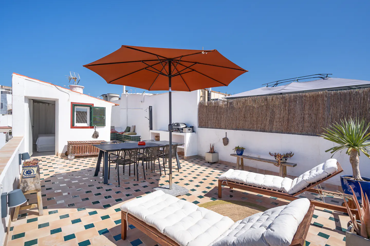 Rooftop patio with lounge chairs, dining table, and orange umbrella under a clear blue sky. White walls and patterned tile flooring.