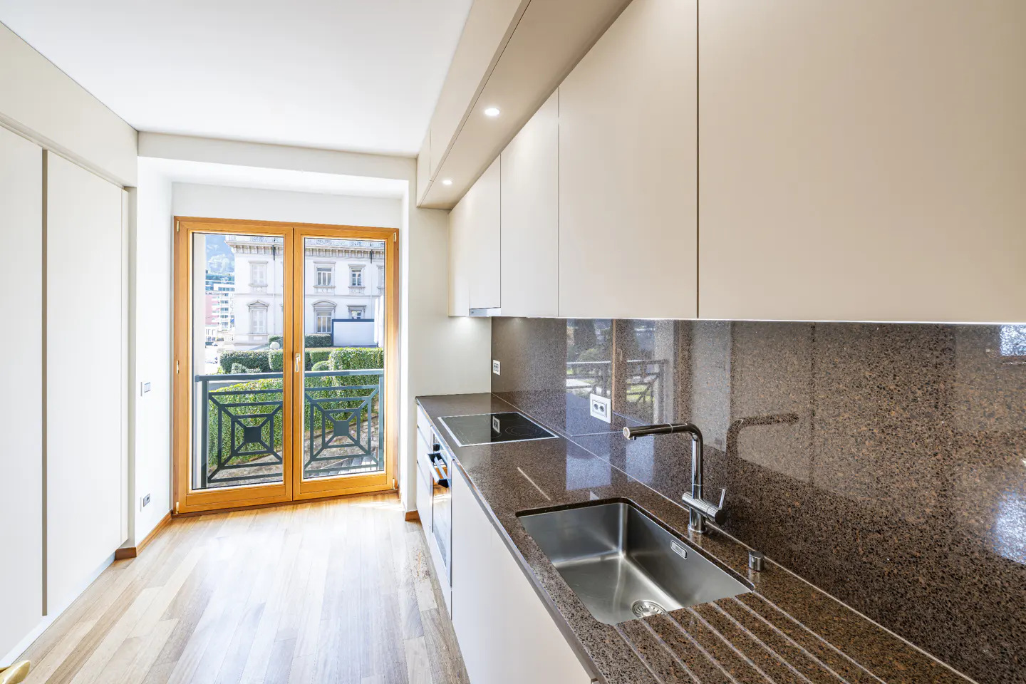 Modern kitchen with light wood floors, beige cabinets, and brown granite countertops. A double door leads to a balcony with a view of a building.