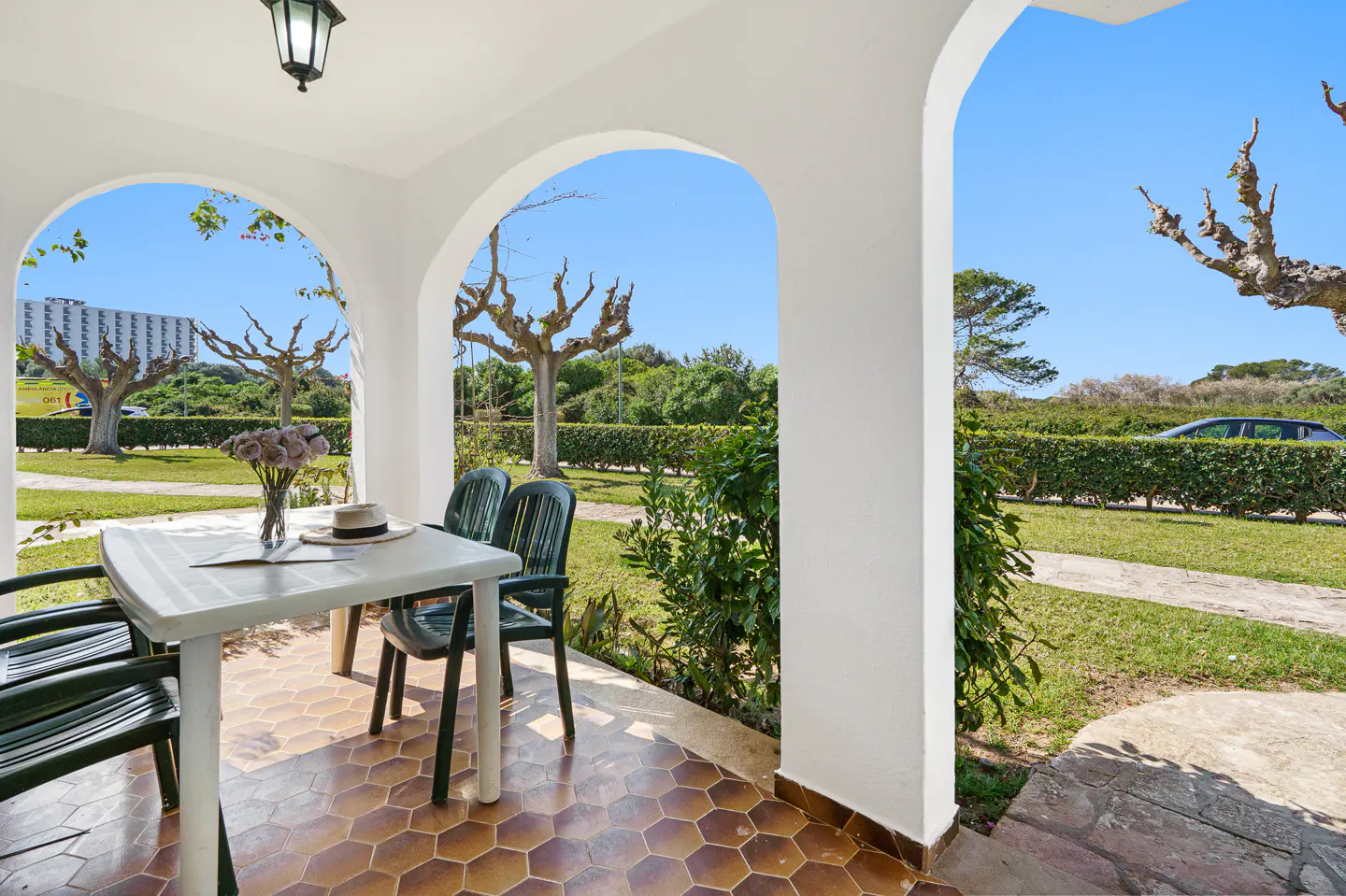 Covered patio with white arches, a white table, and green chairs. A vase of flowers and a hat sit on the table. Outside is a green lawn and blue sky.