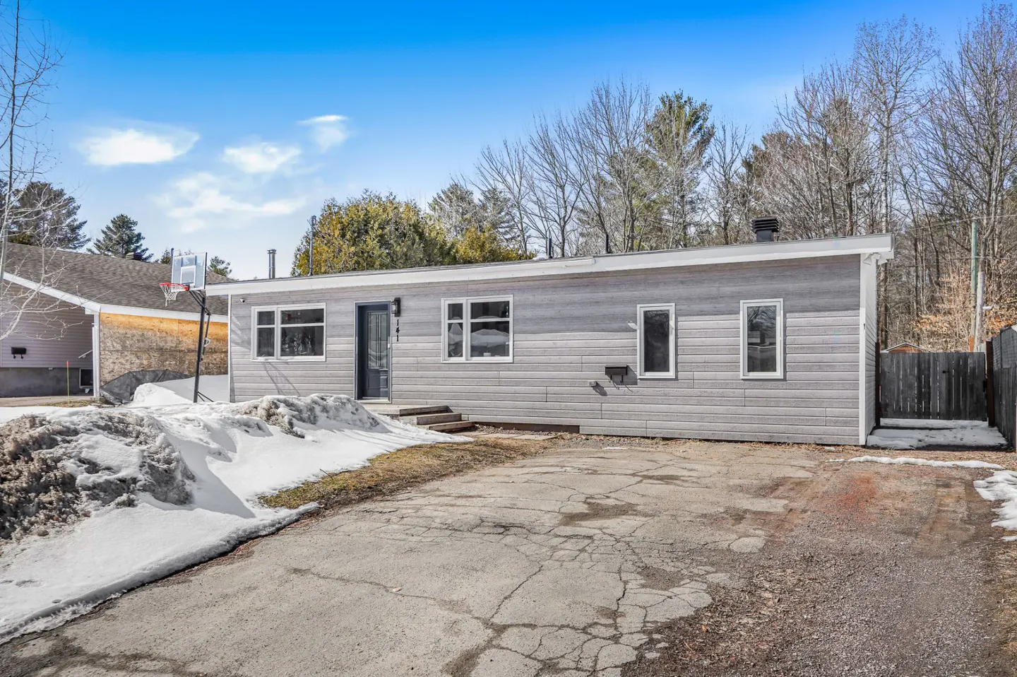 Exterior of a one-story gray house with a black door, windows, and a cracked driveway. Snow is on the lawn. Blue sky.