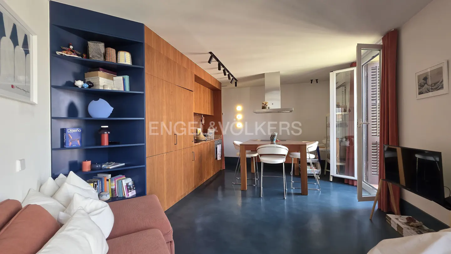 A modern apartment interior with blue floors, wood cabinets, and a blue bookshelf. A dining table with white chairs sits near an open balcony door.