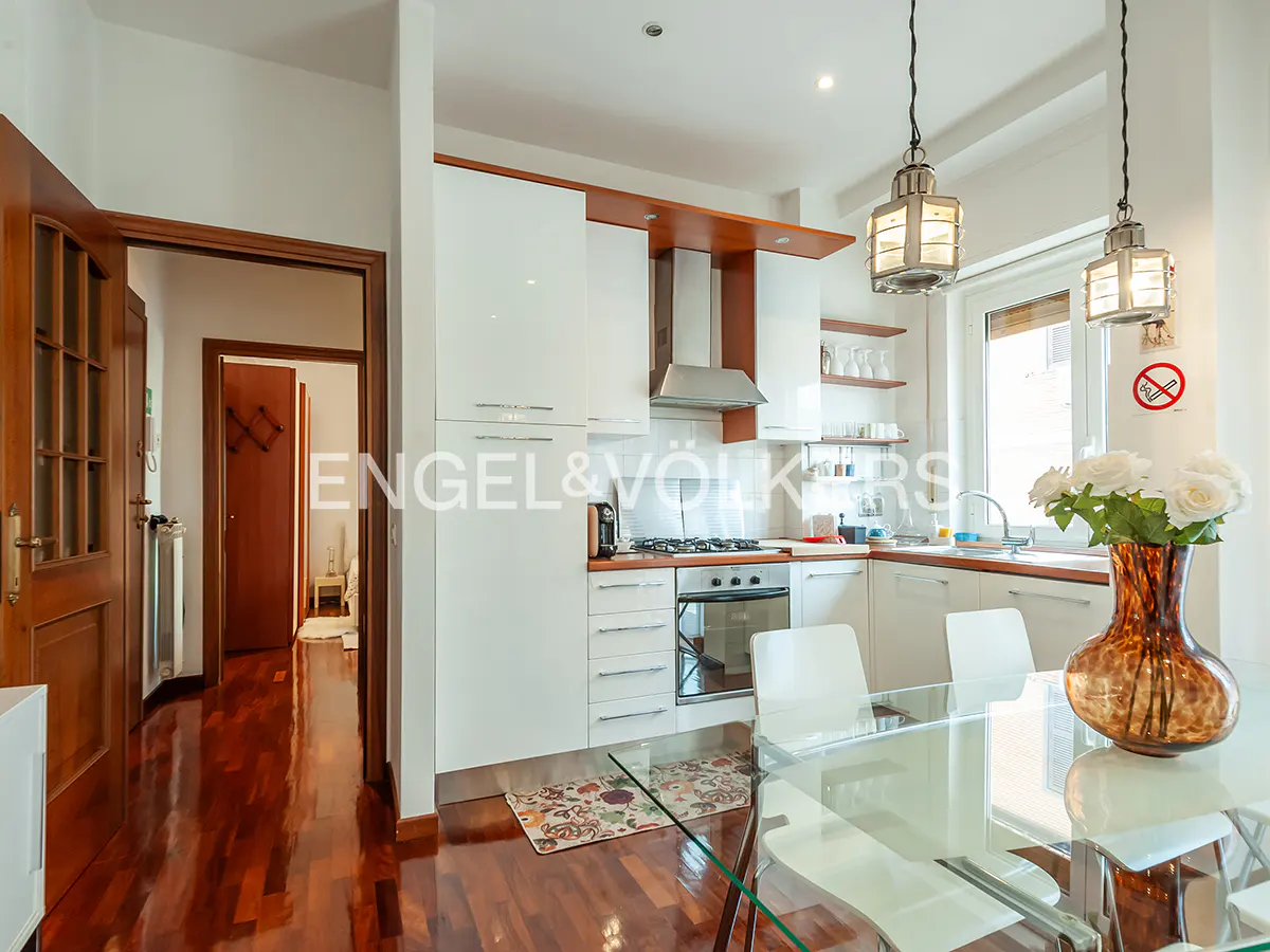 A bright kitchen with white cabinets, stainless steel appliances, and a glass dining table with white chairs. A vase of white roses sits on the table.