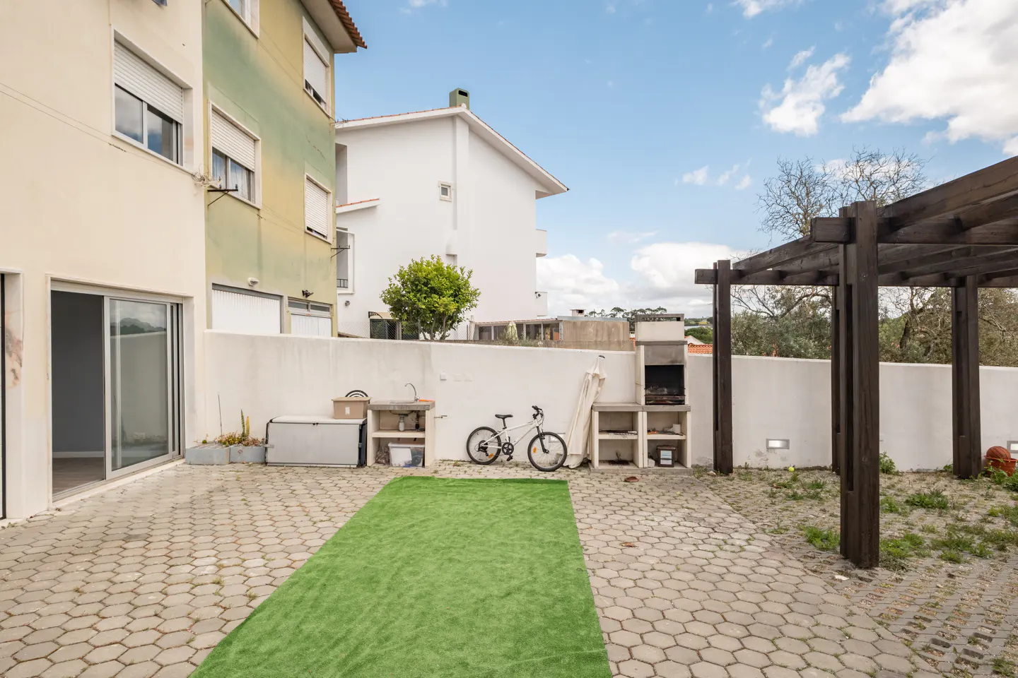 A backyard with a stone patio, a green artificial turf, a grill, and a bicycle against a white wall.